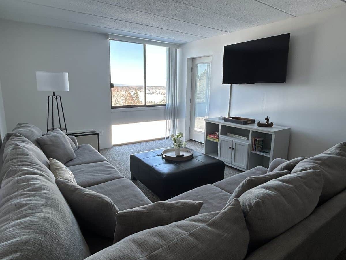 A modern living room with a sectional sofa, a coffee table, a flat-screen TV mounted on the wall, and a bookshelf.
