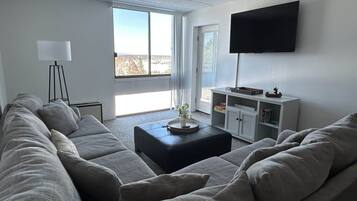 A modern living room with a sectional sofa, a coffee table, a flat-screen TV mounted on the wall, and a bookshelf.