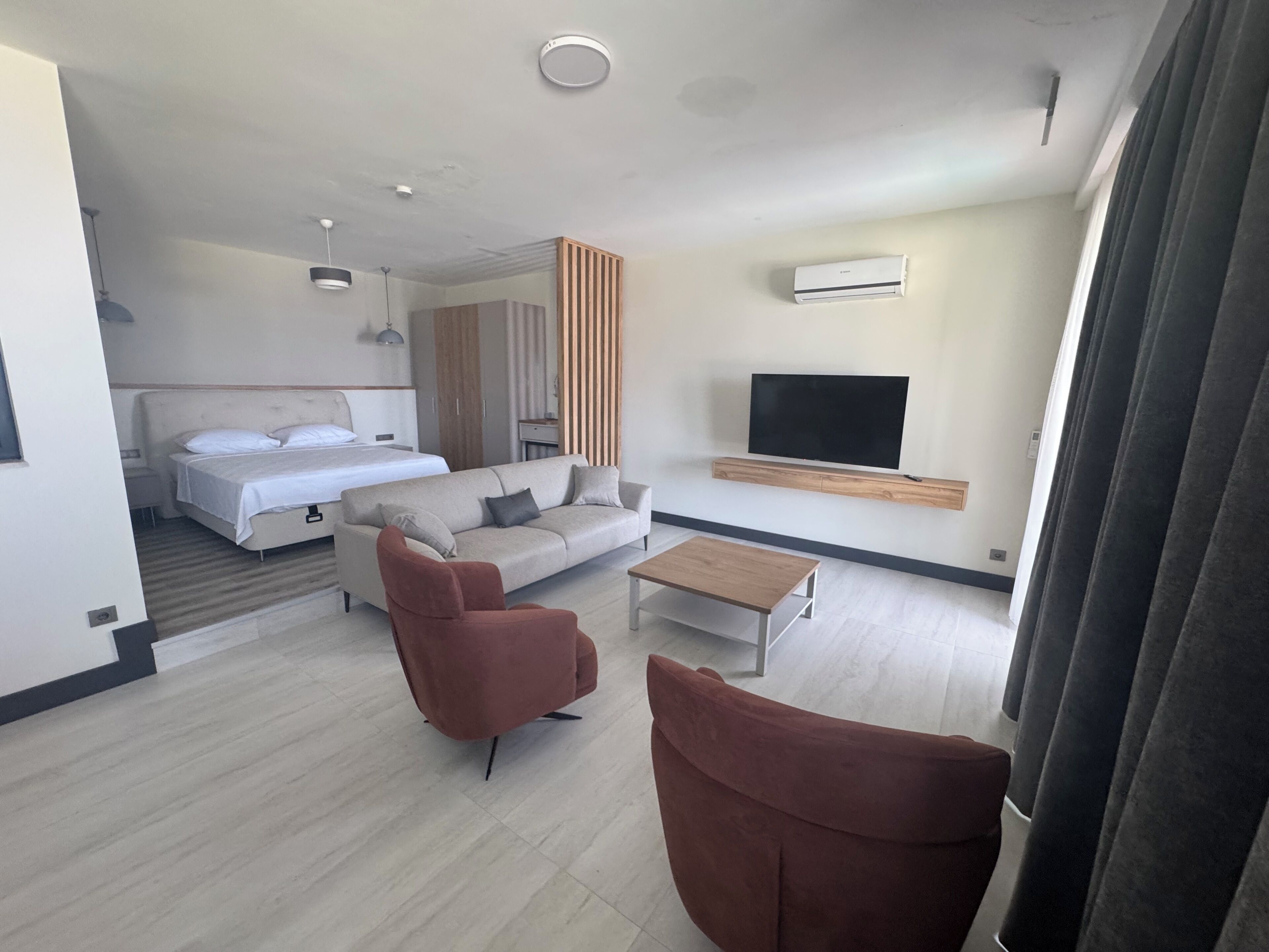 A modern hotel room with a bed, sofa, two armchairs, a coffee table, a TV, and an air conditioning unit.