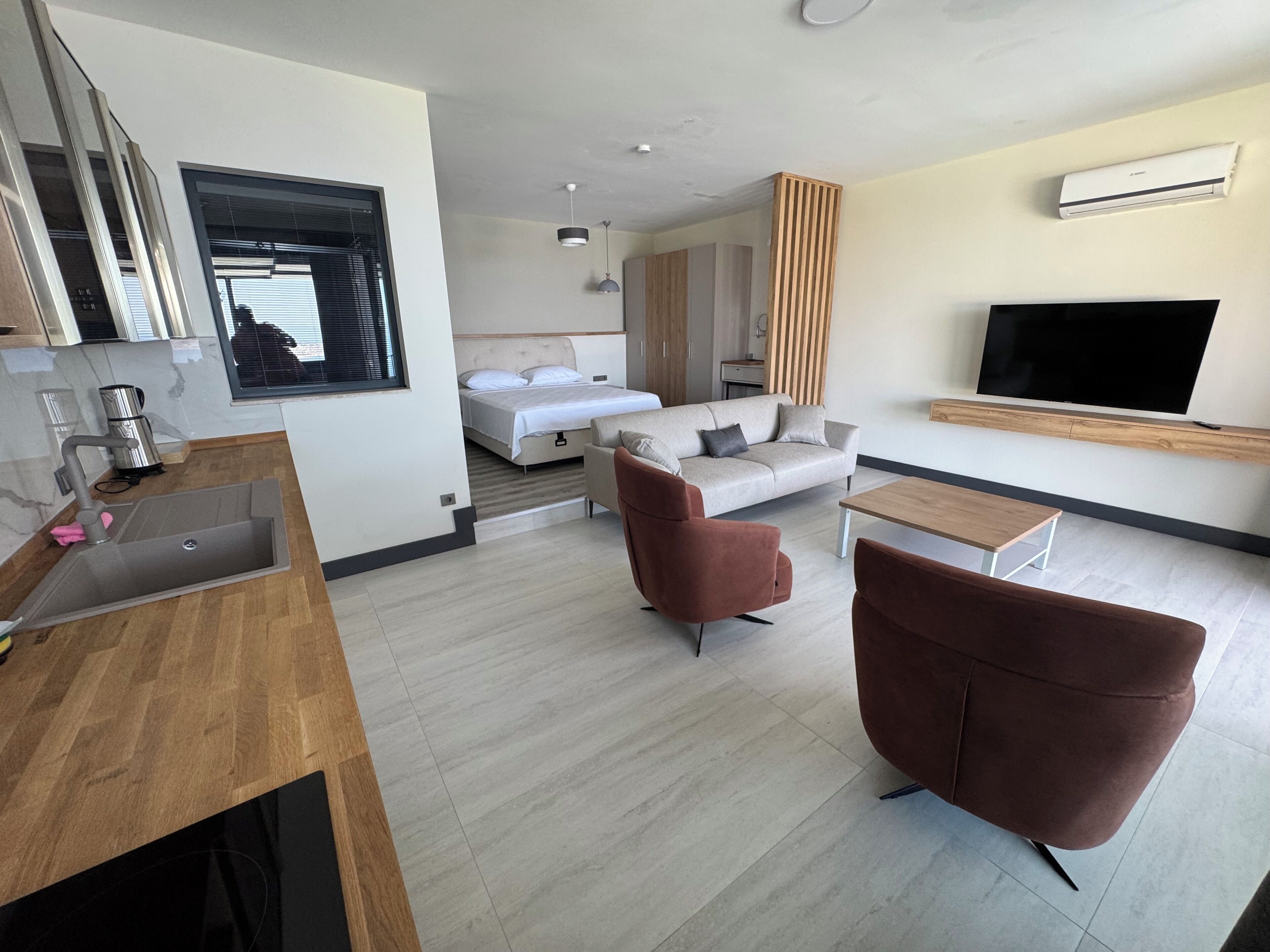 A modern hotel room with a kitchenette, a bed, a sofa, a TV, and a wooden floor.