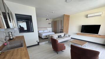 A modern hotel room with a kitchenette, a bed, a sofa, a TV, and a wooden floor.
