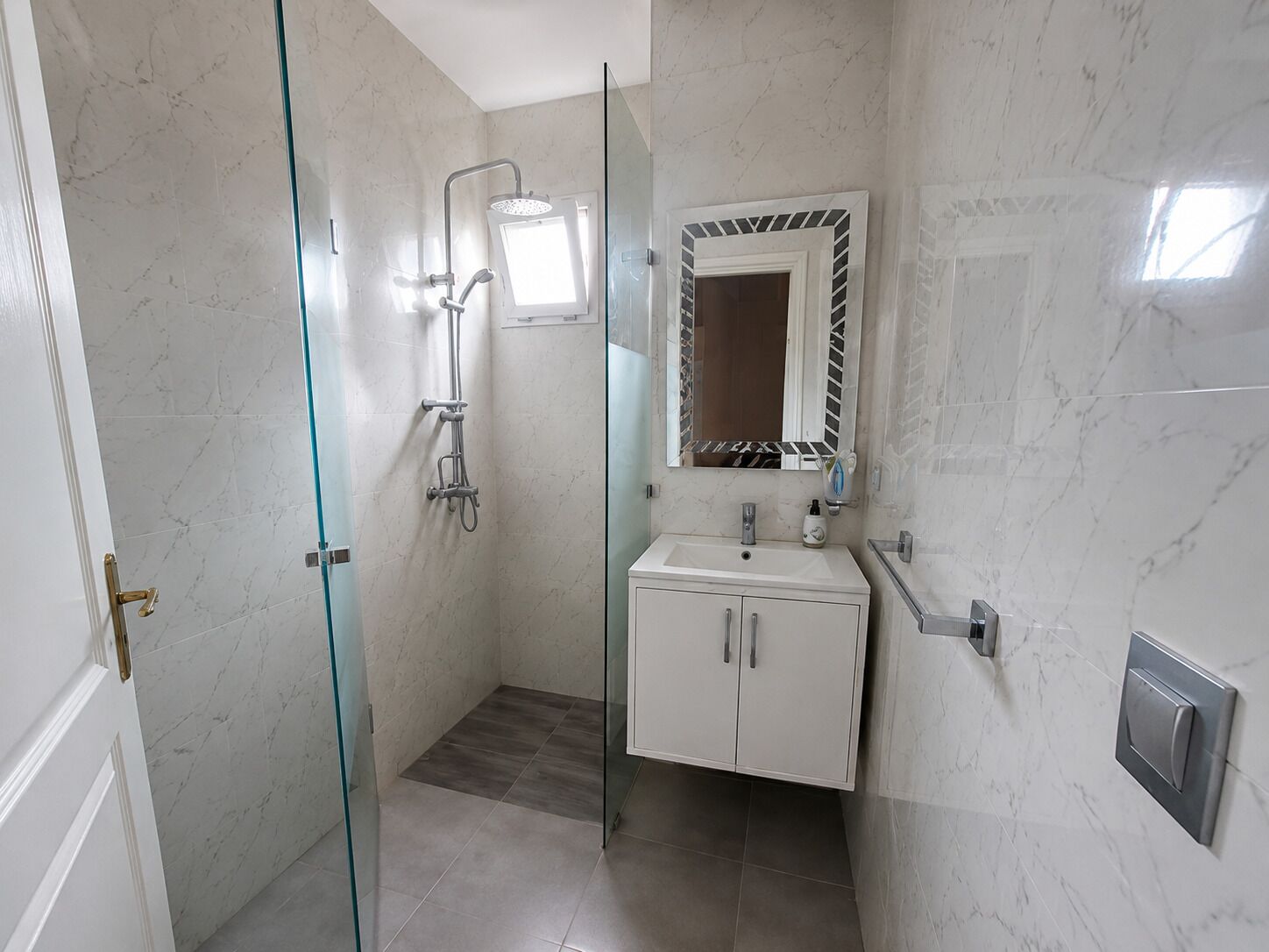 A bathroom with a glass-enclosed shower, a white sink, and a mirror with a decorative frame.