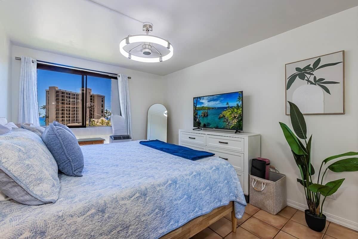 A bedroom with a bed, a television, a mirror, and a window with a view of buildings.