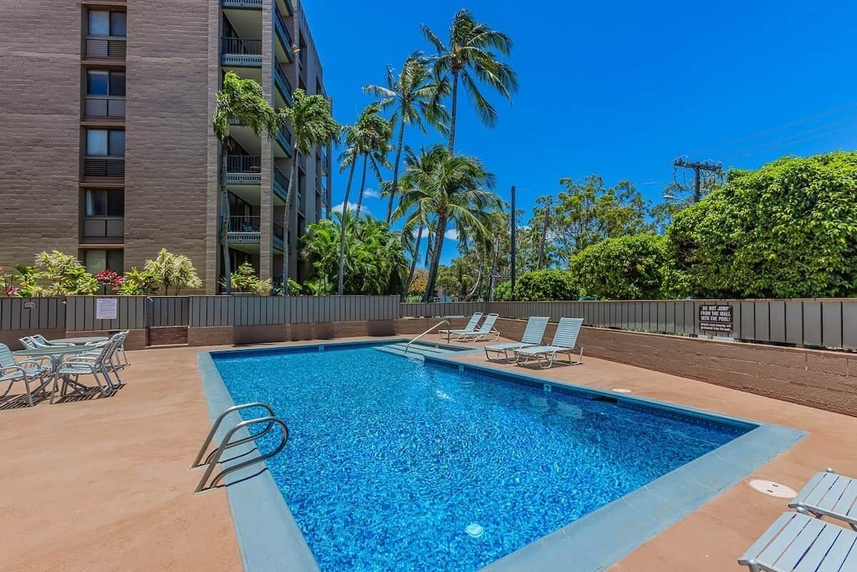 A rectangular swimming pool with a metal ladder, surrounded by lounge chairs and palm trees.