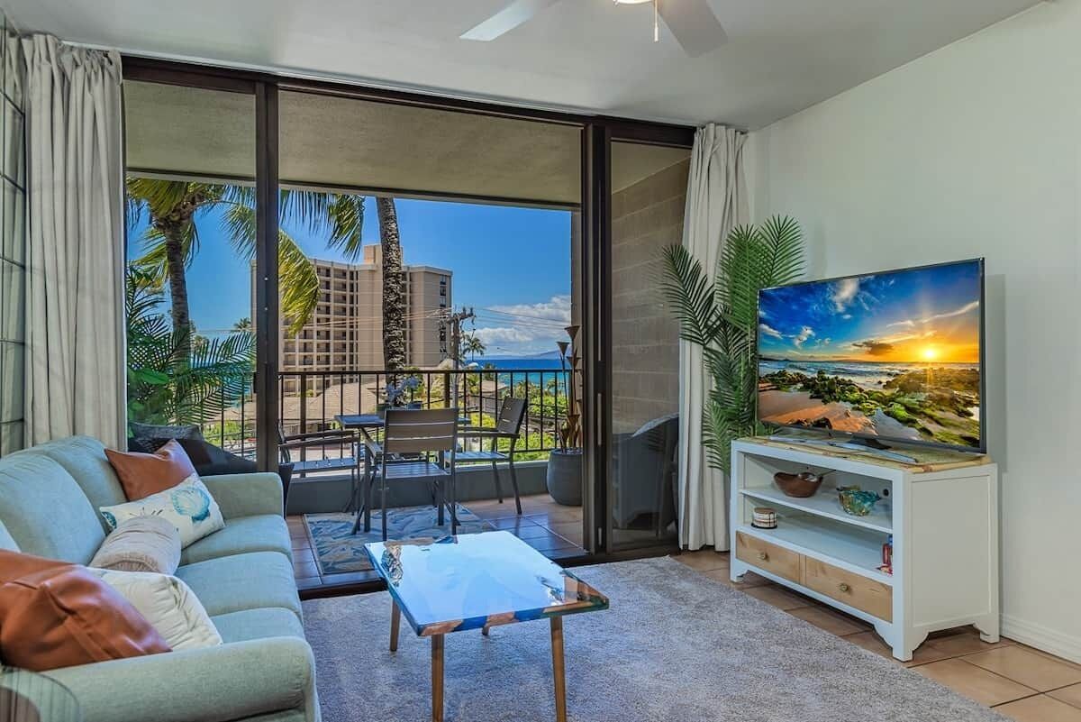 A living room with a sofa, coffee table, TV, and a balcony with a view of palm trees and buildings.