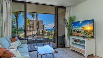 A living room with a sofa, coffee table, TV, and a balcony with a view of palm trees and buildings.