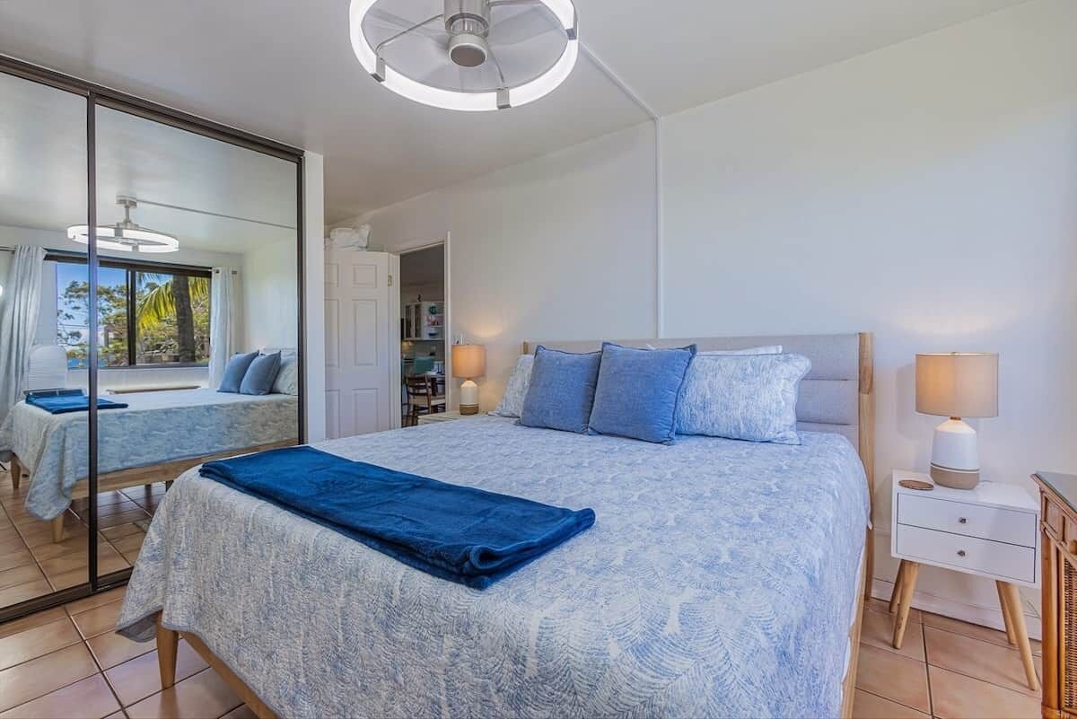 A bedroom with a large bed, a bedside table, a mirror, and a view of the outdoors.
