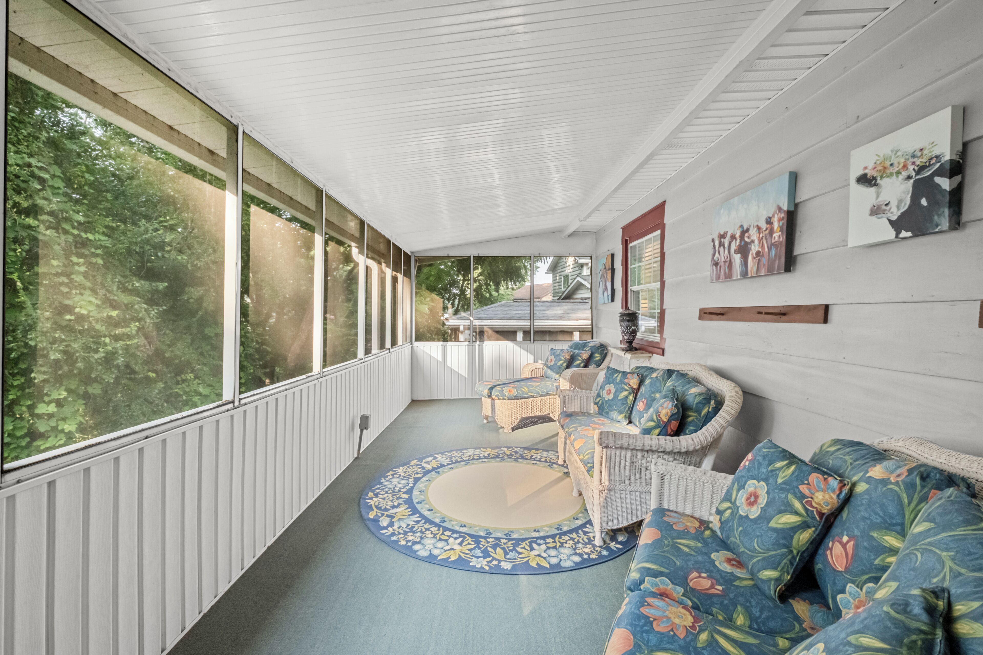 A covered porch with wicker furniture, a round rug, and artwork on the walls.