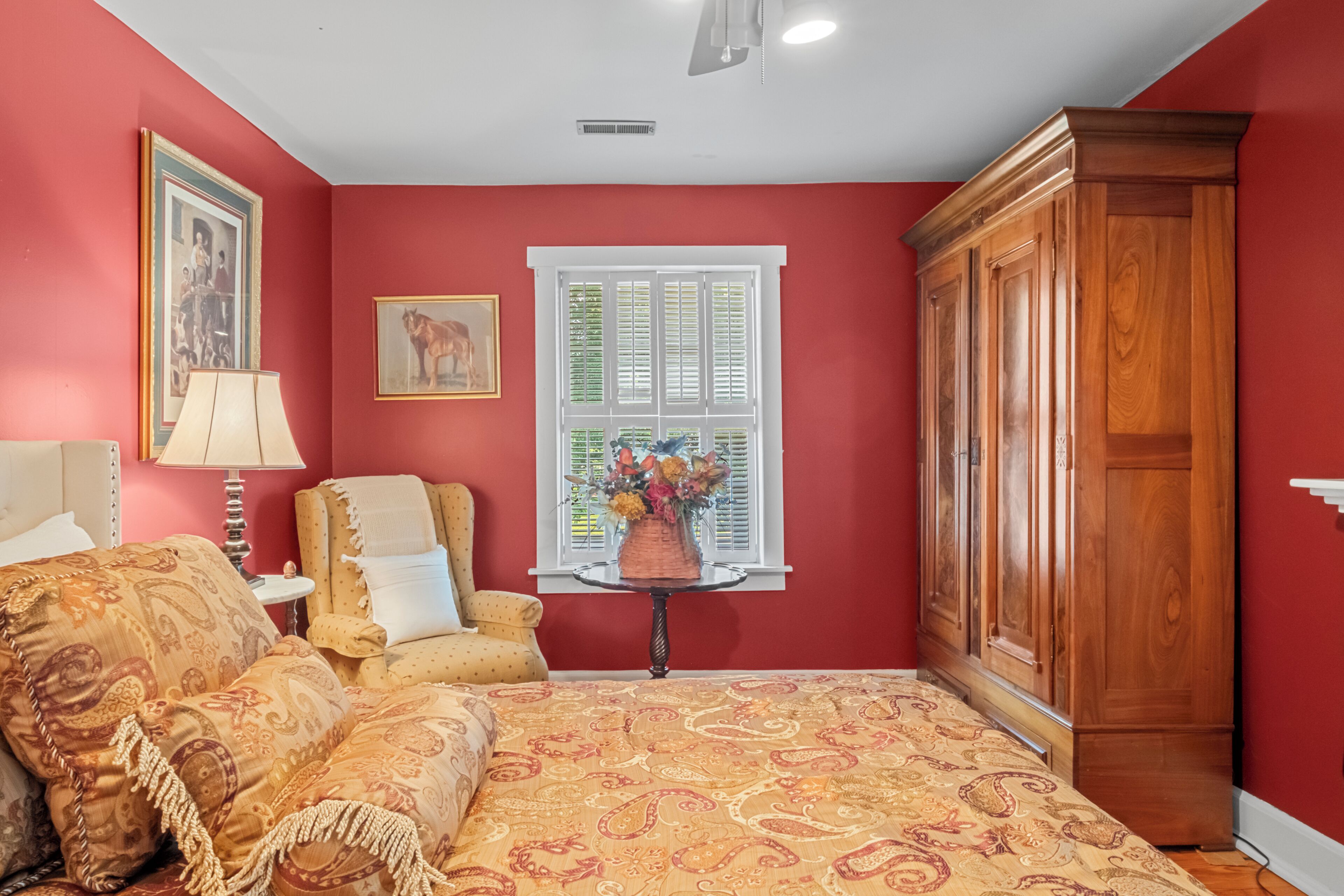 A room with a bed, a chair, a table with flowers, a window with shutters, and a wardrobe.
