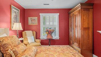 A room with a bed, a chair, a table with flowers, a window with shutters, and a wardrobe.