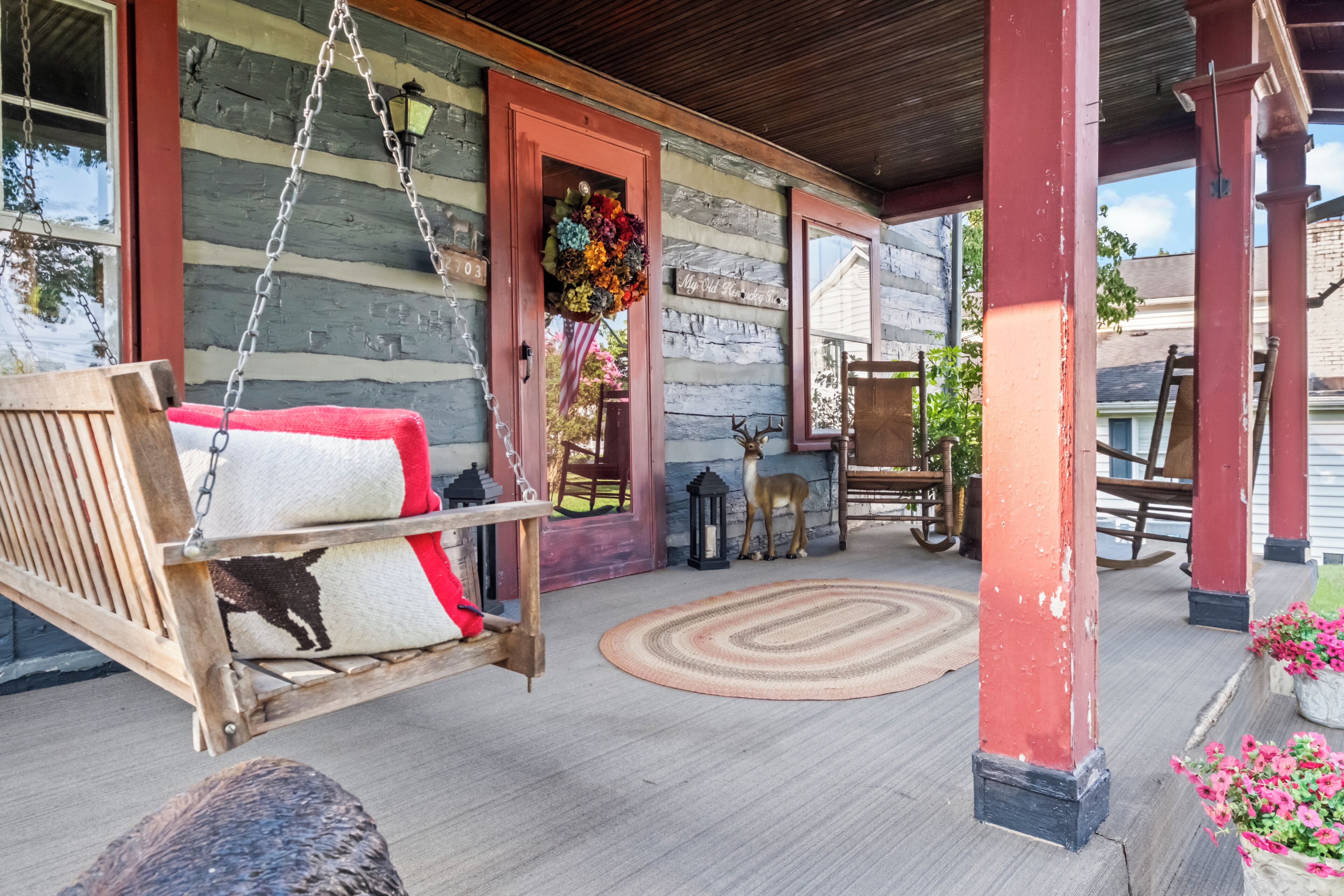 A porch with a swing, a wooden bench, a decorative dog statue, a wooden door, and a hanging flower arrangement.