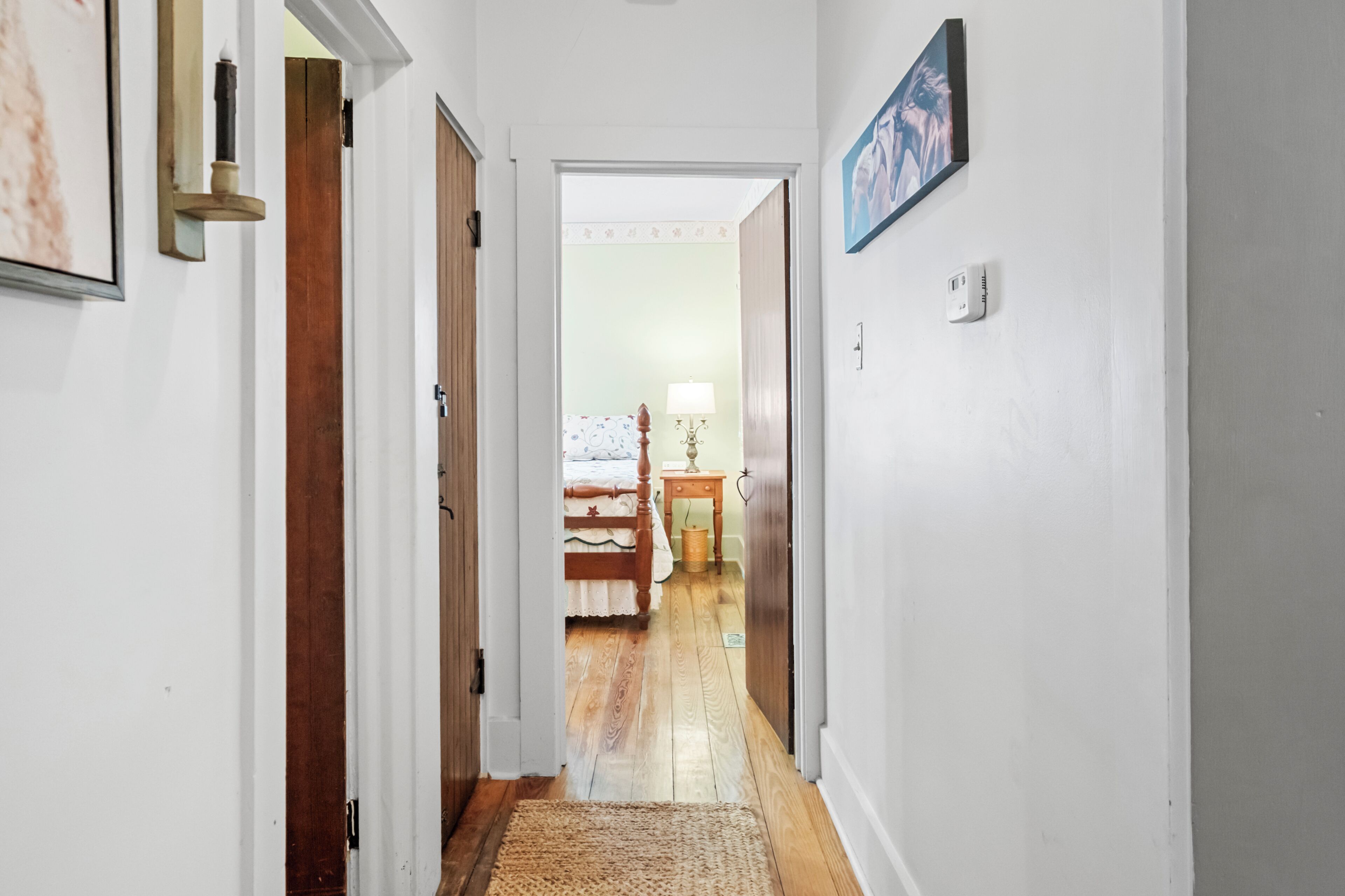 A hallway with a wooden floor, a rug, a bed with a white and blue patterned bedspread, and a nightstand with a lamp.