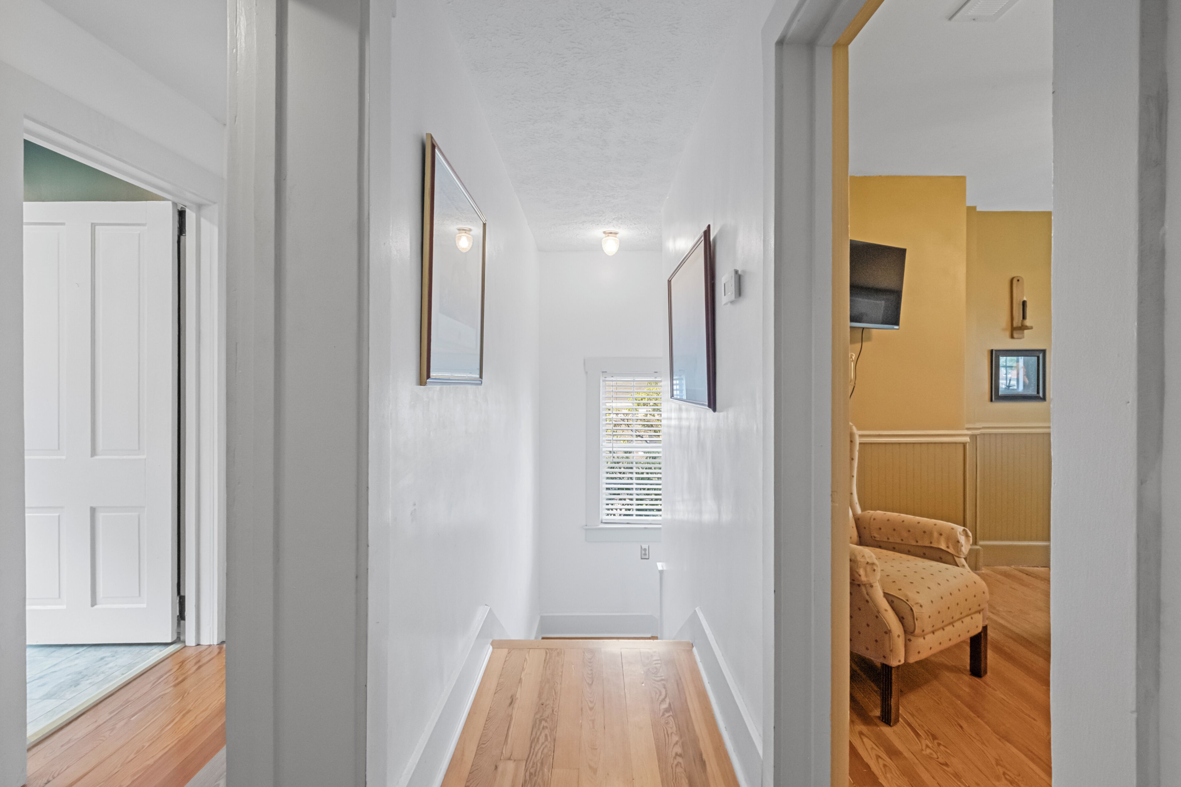 Un couloir avec un plancher en bois, des murs blancs et une porte au fond. Deux miroirs encadrés sont accrochés au mur, et il y a une fenêtre avec des stores.