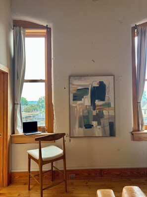 Ein Zimmer mit Holzboden, Schreibtisch mit Laptop, Stuhl, gerahmtes abstraktes Gemälde und Fenster mit Vorhängen.