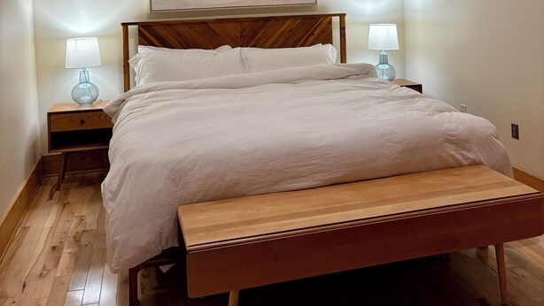 A bedroom with a bed, wooden headboard, two bedside tables with lamps, a bench, and a painting on the wall. - Old City 2BR Condo close to Downtown and UTK: excellent walkablity (Knoxville)