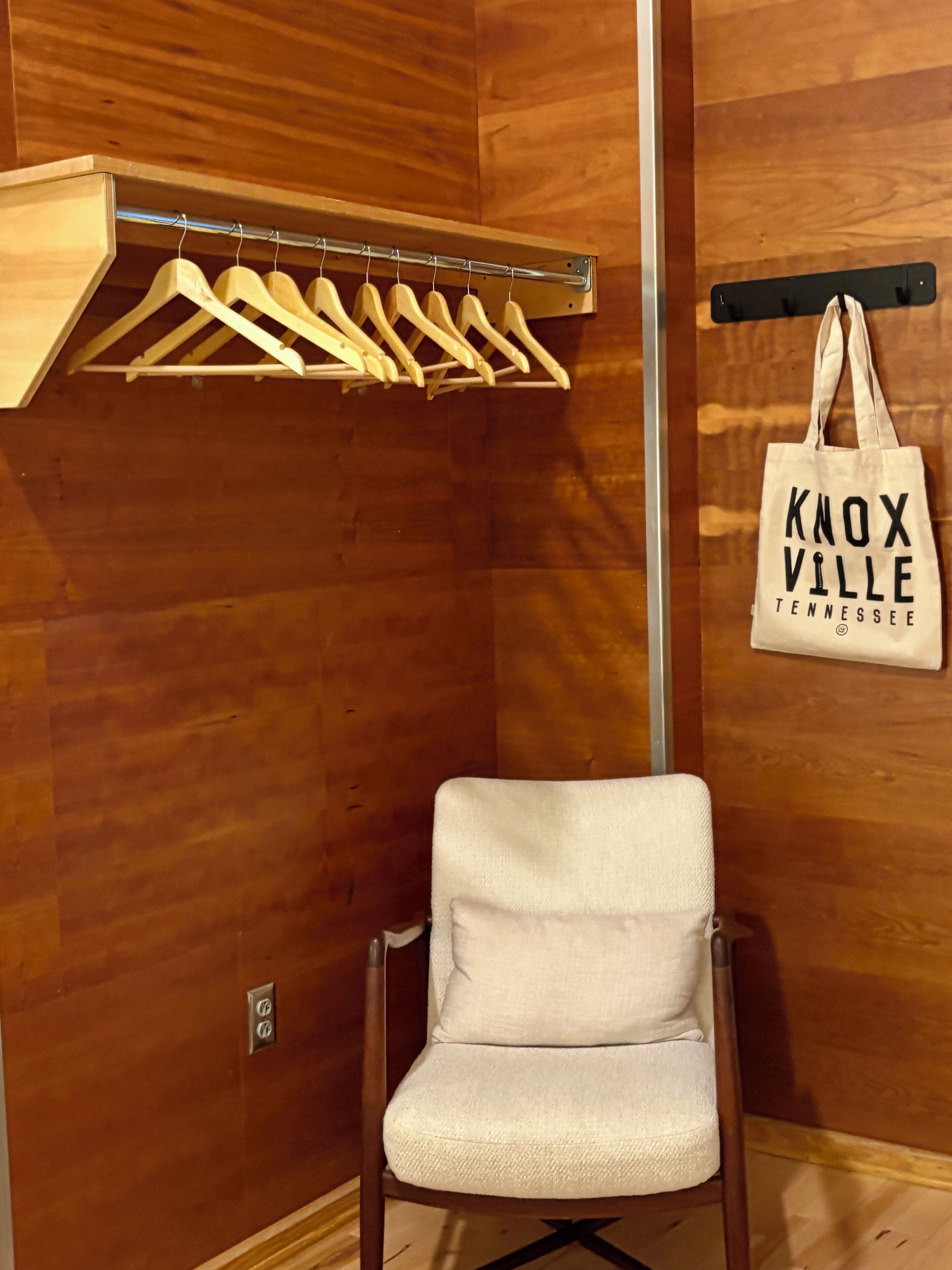Une pièce avec un mur en bois, une chaise blanche, des cintres en bois sur un portant en métal et un sac fourre-tout avec l’inscription « Knoxville, Tennessee ».