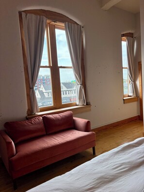 Ein Zimmer mit einem roten Sofa, zwei mit Vorhängen versehenen Fenstern, Holzboden und Blick auf Gebäude.