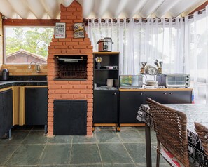 A kitchen with a brick oven, a sink, a stove, and a dining area with a wicker chair. - Cabana Mata Verde Pool/Lake - Tomorrowland (Itu)