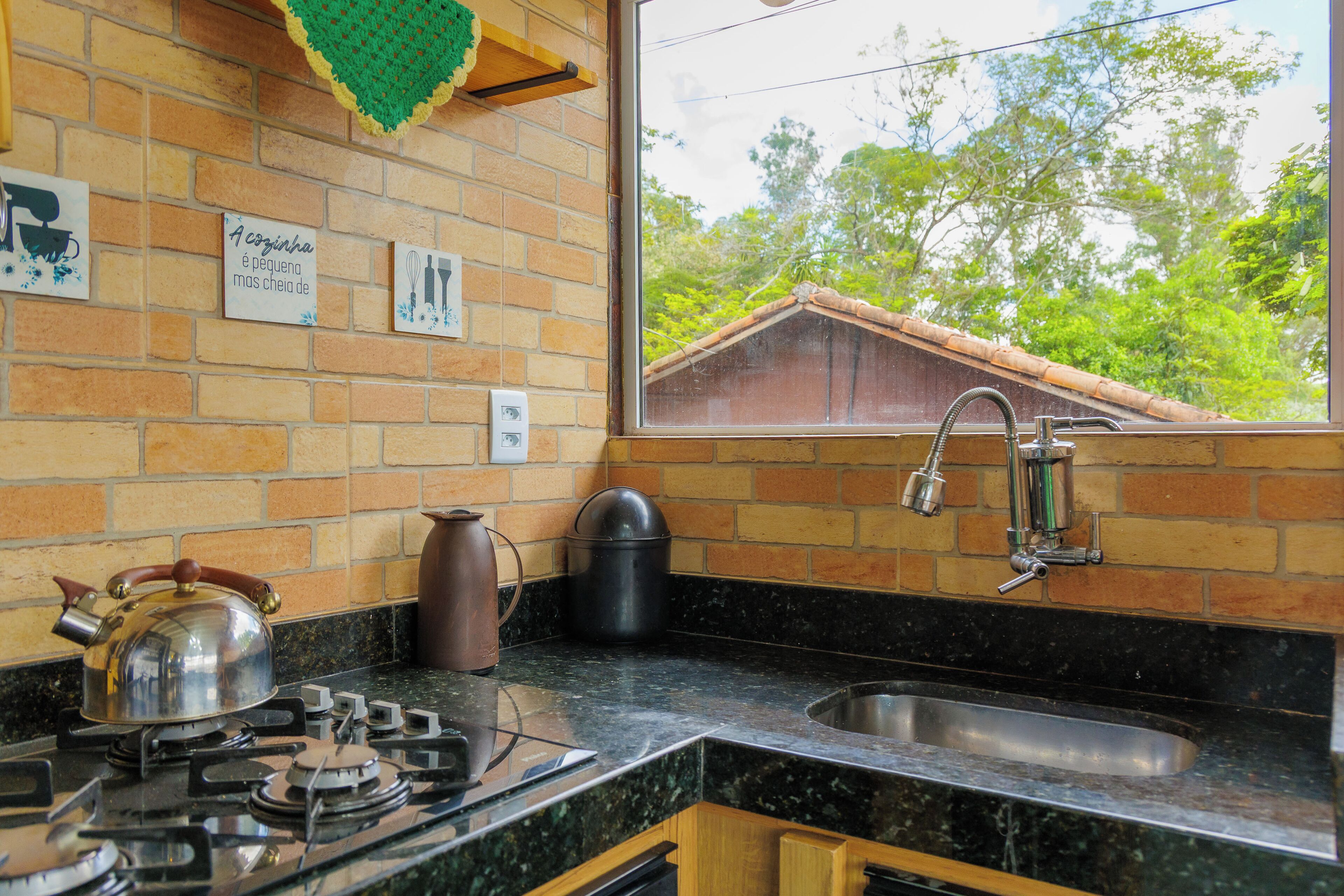 A kitchen with a gas stove, a kettle, a sink, and a window with a view of trees.