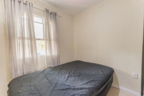 Um quarto pequeno e simples, com uma cama de solteiro, uma janela com cortinas leves e uma parede sem adornos.