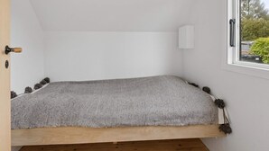 Ein kleiner, gemütlicher Raum mit schräg versiegelter Decke, einem Einzelbett mit gestreiftem Bettbezug, einer weißen Wandleuchte und einem Fenster mit Blick auf Bäume und parkende Autos.