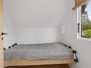 Ein kleiner, gemütlicher Raum mit schräg versiegelter Decke, einem Einzelbett mit gestreiftem Bettbezug, einer weißen Wandleuchte und einem Fenster mit Blick auf Bäume und parkende Autos.