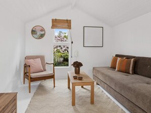 A cozy living room with a wooden chair, a brown sofa, a small wooden table, and a window with a view of cherry blossoms.