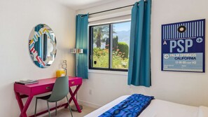 A hotel room with a desk, chair, bed, window with curtains, a mirror, and a wall poster. - Nicola Oasis Resort-Style Palm Springs Oasis | Saltwater Pool, Spa & Mountain Views (Palm Springs)