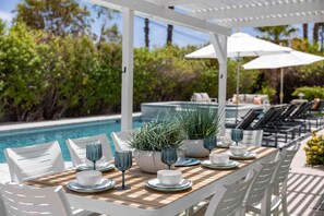 A poolside dining area with white chairs, a wooden table, and umbrellas. - Nicola Oasis Resort-Style Palm Springs Oasis | Saltwater Pool, Spa & Mountain Views (Palm Springs)