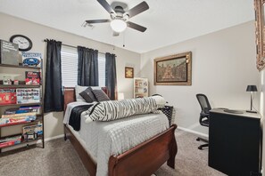 Um quarto com uma cama, escrivaninha, cadeira, ventilador de teto, relógio e estante de livros.