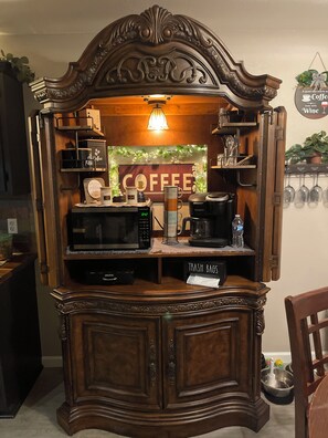 Estação de café com placa indicando 'COFFEE', uma cafeteira Keurig, uma lixeira e um relógio marcando 6:42.