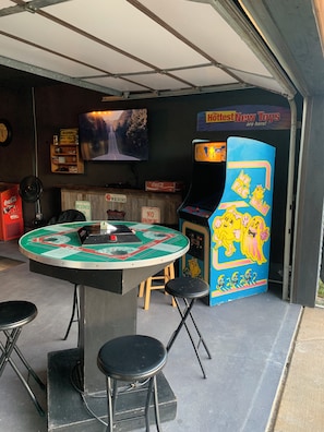 An indoor arcade with a vintage pinball machine, a jukebox, and a television screen displaying a road scene. - Cozy Getaway, an ultimate Tulsa suburb retreat and a backyard oasis. (Tulsa)