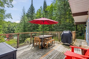 A wooden deck with a table and chairs, a red umbrella, and a barbecue grill. - Muskoka River Cottage - Kayaks, Firepit, BBQ, Wifi (Bracebridge)