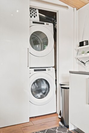 A compact laundry room with a front-loading washer and dryer, a trash bin, and a small shelf with a radio. - Muskoka River Cottage - Kayaks, Firepit, BBQ, Wifi (Bracebridge)