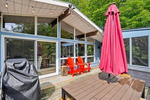 A modern house with large glass windows, a wooden deck, outdoor seating, and a covered patio area. - Muskoka River Cottage - Kayaks, Firepit, BBQ, Wifi (Bracebridge)
