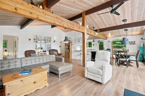 A spacious living area with a large sectional sofa, a white armchair, and a wooden coffee table. There is a kitchen area with a dining table and chairs visible in the background. - Muskoka River Cottage - Kayaks, Firepit, BBQ, Wifi (Bracebridge)
