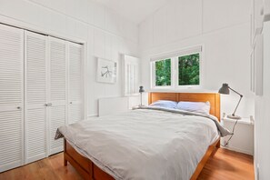 Een slaapkamer met een bed, nachtkastje, raam met uitzicht op bomen en kast met witte luiken.