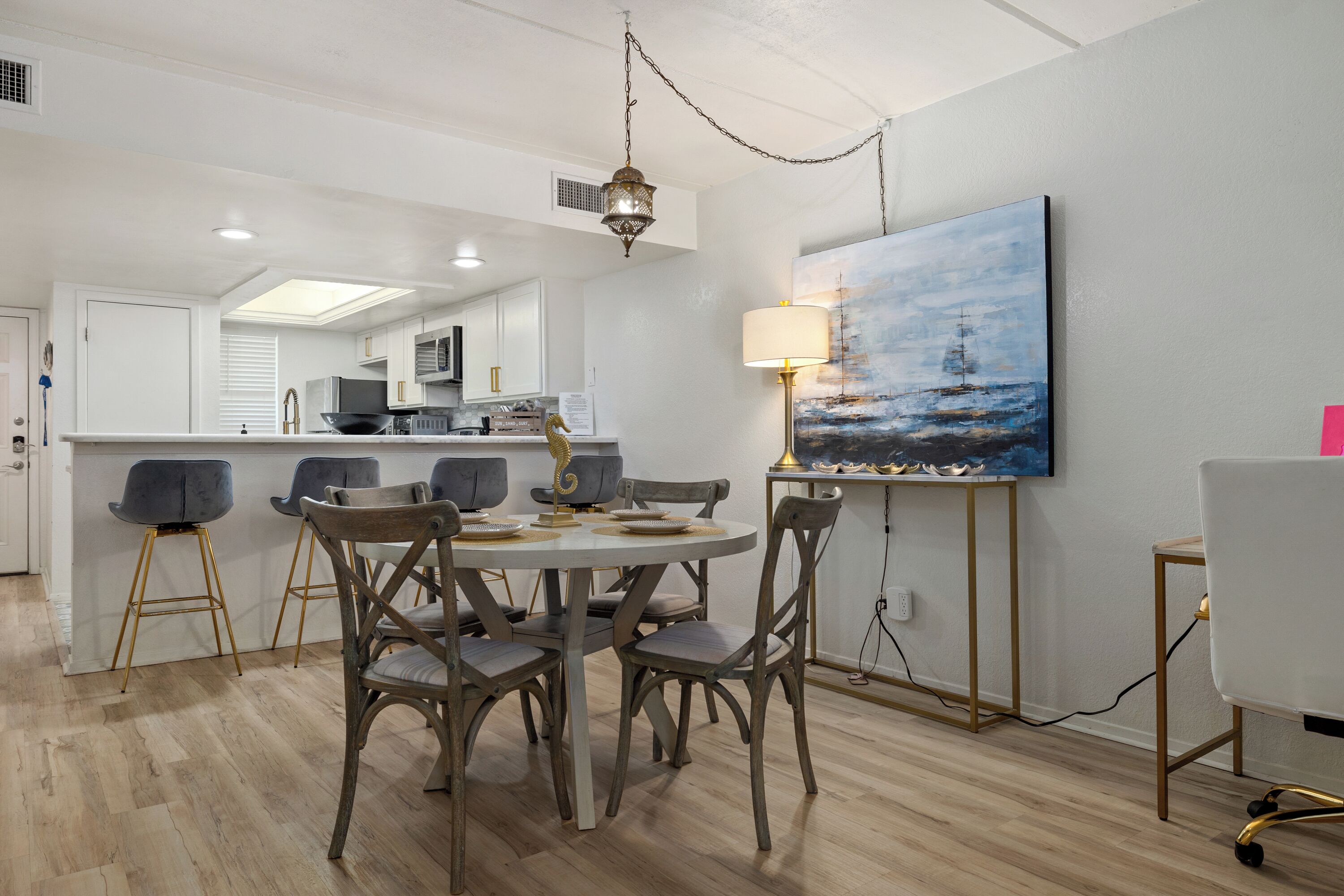 Une cuisine moderne avec des meubles blancs, un coin repas avec une table ronde et des chaises, et un tableau représentant un paysage marin accroché au mur.