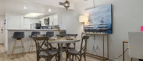Une cuisine moderne avec des meubles blancs, un coin repas avec une table ronde et des chaises, et un tableau représentant un paysage marin accroché au mur.