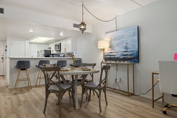 A modern kitchen with white cabinets, a dining area with a round table and chairs, and a painting of a seascape on the wall. - Seas the day! Just a 5 minute walk to the beach! 2bd 2 bath sleeps 8 (South Padre Island)