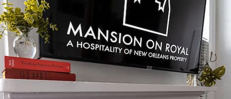 Un televisor de pantalla plana montado en una repisa blanca sobre una chimenea, con un logotipo de corona decorativa y el texto 'MANSION ON ROYAL A HOSPITALITY OF NEW ORLEANS PROPERTY'.