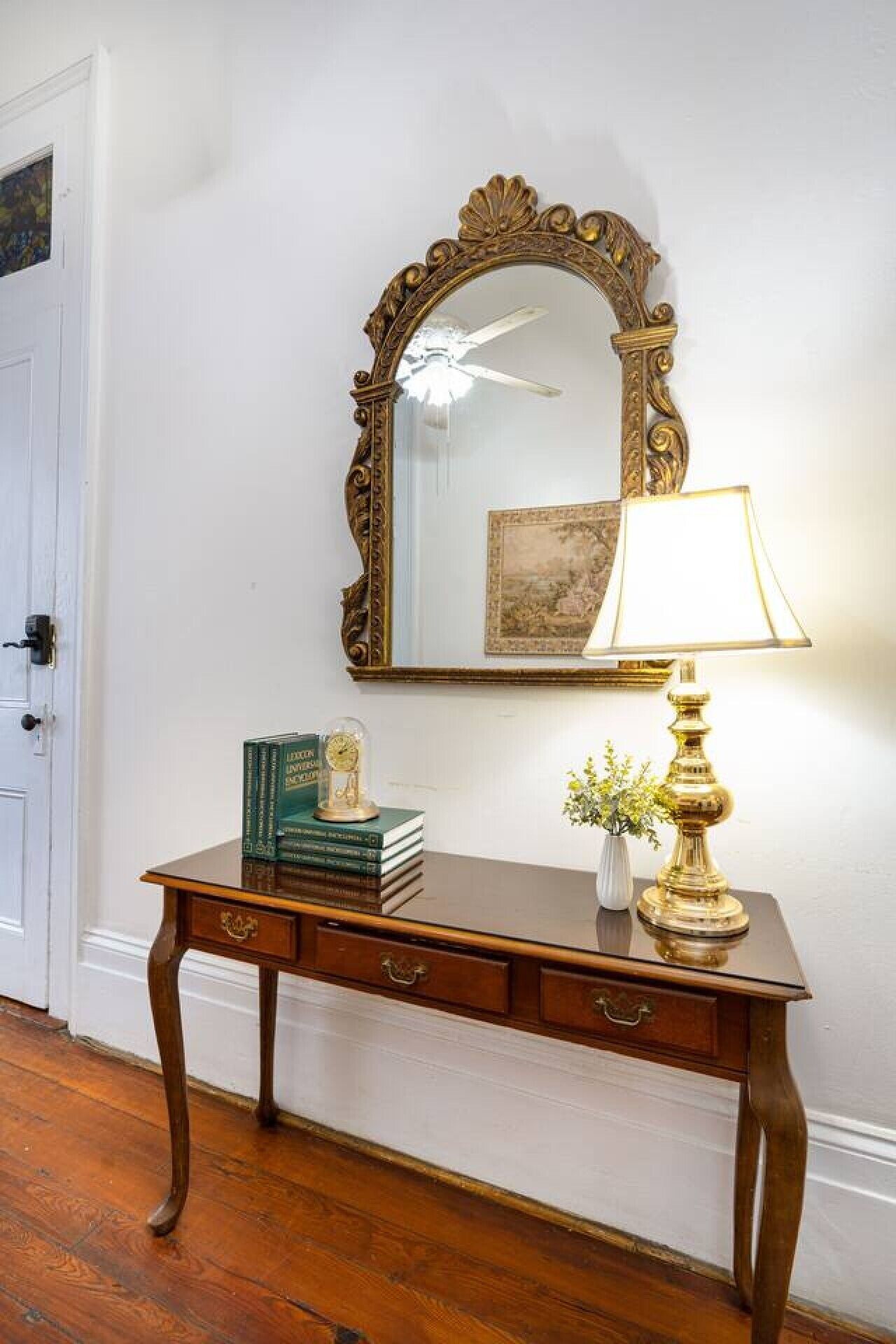 Une console en bois, ornée d’un miroir, de livres, d’une horloge et d’une lampe, est placée contre un mur blanc.