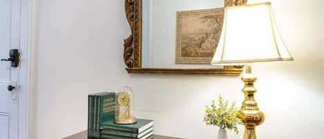 A wooden console table with a mirror, books, a clock, and a lamp against a white wall.