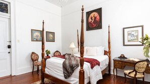 Uma cama com dossel, um gato, uma escrivaninha com cadeira e quadros nas paredes.