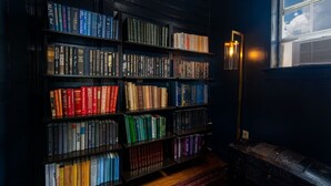 Uma estante cheia de livros, um abajur e uma janela com vista para o céu.