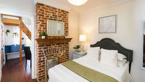 A bedroom with a brick fireplace, a bed with white bedding, a nightstand with a lamp, and a framed picture on the wall. - Historic Carriage House | Walk to Quarter & Music (New Orleans)