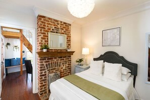 Un dormitorio con chimenea de ladrillo, una cama con ropa de cama blanca, una mesita de noche con lámpara y un cuadro en la pared.