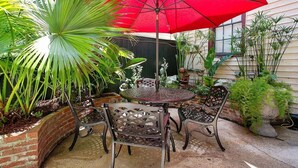 An outdoor patio with a round metal table and chairs, a red umbrella, and various potted plants. - Historic Carriage House | Walk to Quarter & Music (New Orleans)