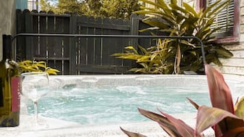 A hot tub with a wooden deck, surrounded by plants and a fence.