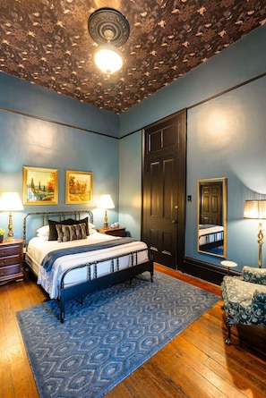 Et soveværelse med seng, natborde, et spejl, en dør og et mønstret tæppe.